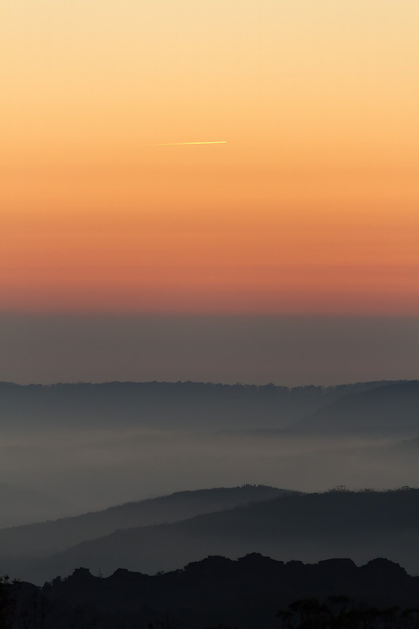 A contrail catching the dawn above receding misty hills at Mount Tomah on the Bells Line of Road in NSW, Australia.