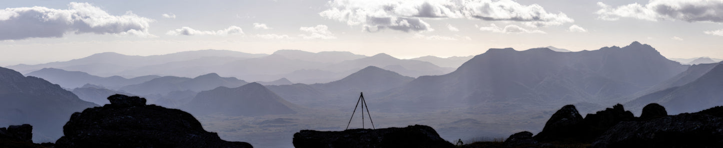 An amazing panorama capturing the essence of Out Under The Sky. My tripod is silhouetted against a back drop of mountain ranges fading to the horizon in silver afternoon light which lights the clouds from above. One of my favourites. Taken from atop the Western Arthurs in Tasmania, Australia.